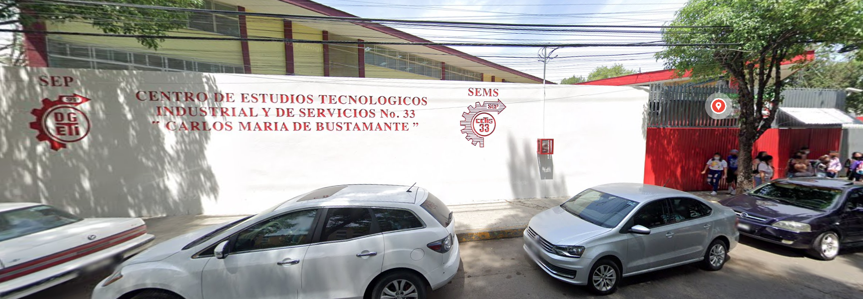 Fotografía en la que se muestra la fachada del plantel, conformada por un muro de color blanco que en letras grandes rojas dice CENTRO DE ESTUDIOS TECNOLÓGICOS INDUSTRIAL Y DE SERVICIOS NÚMERO 33, CARLOS MARIA DE BUSTAMANTE. Del lado derecho del muro se encuentra un portón rojo por el que se puede ingresar al plantel.
